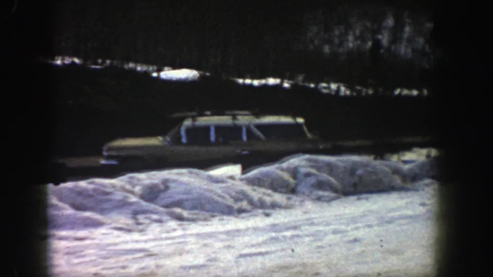 1961 Aspen winter home-movie frame showing a station wagon partly screened by roadside snowbanks and shadowed trees.
