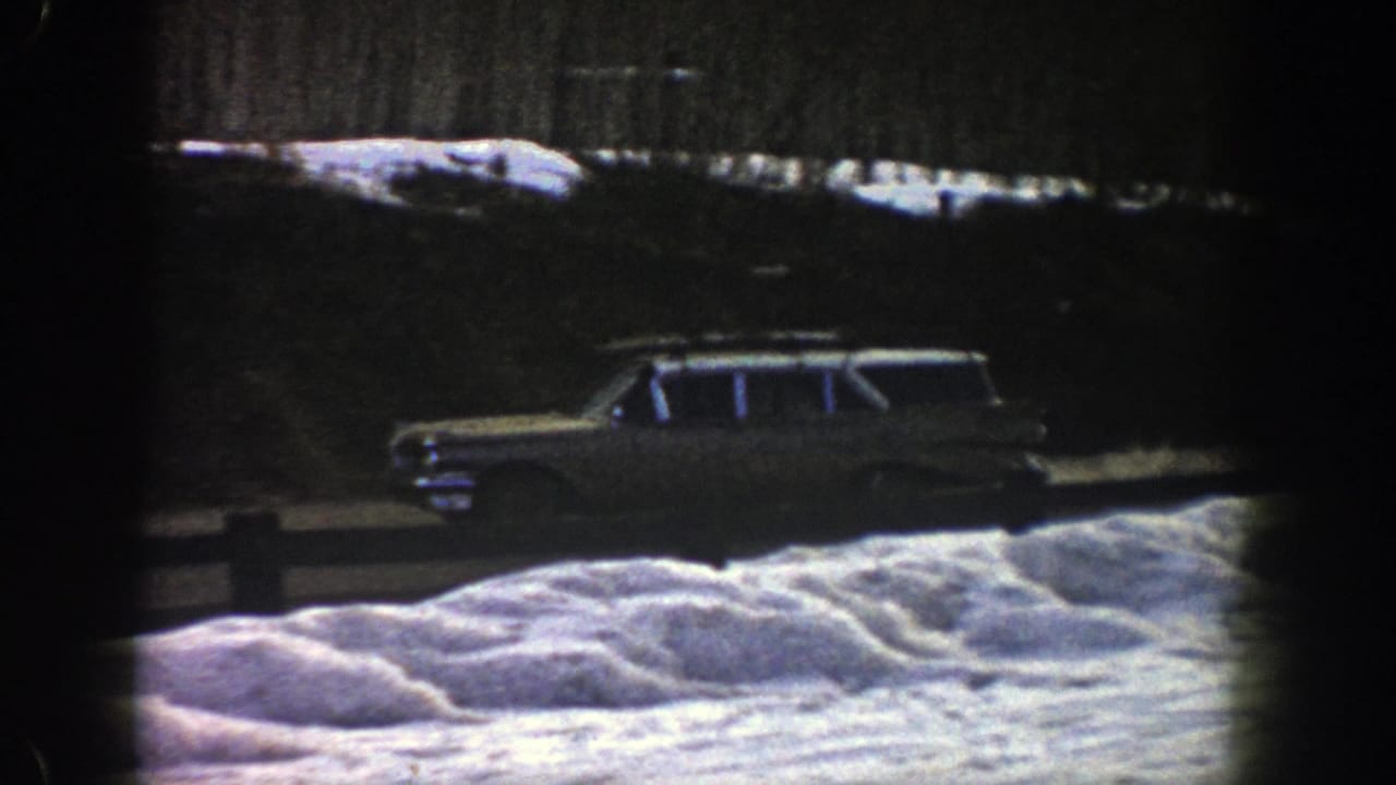 Station wagon with roof rack drives past snowbanks and a wooden roadside rail.