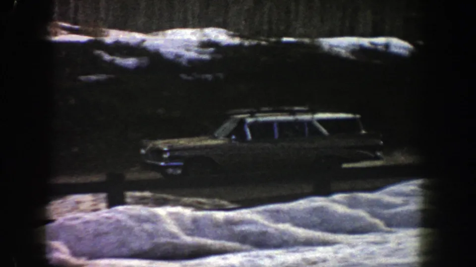 The 1961 Aspen road scene shows the roof-rack station wagon moving past snow and evergreen shadow.