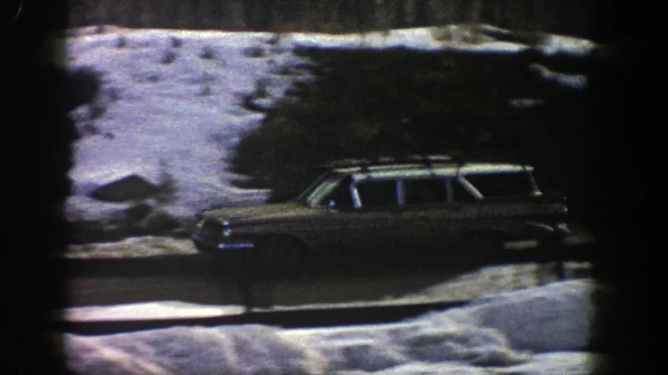 A vintage station wagon travels through a snowy Aspen roadside view, with dark evergreens and exposed pavement visible.