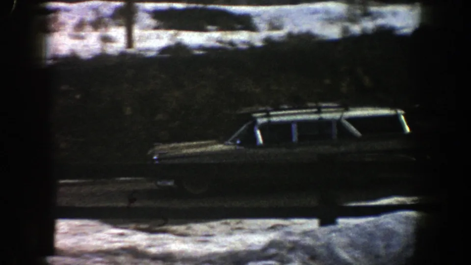 The car continues along the snow-lined Aspen road, framed by a guardrail, dark trees, and 8mm film grain.