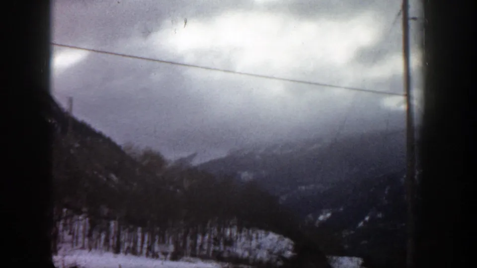 1961 Aspen, Colorado 8mm view of snow-covered trees and layered mountain slopes under a heavy gray sky, with roadside utility infrastructure visible.