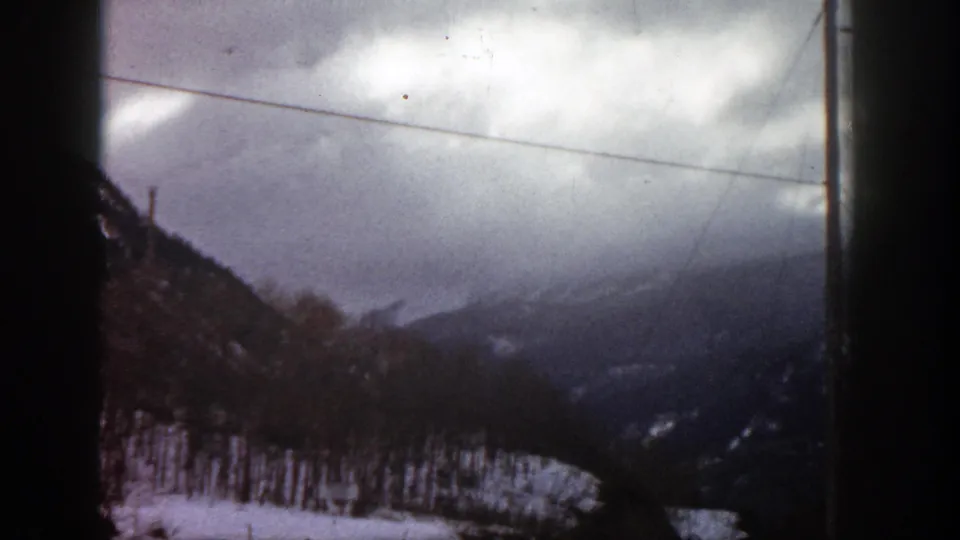 1961 Aspen, Colorado home-movie frame of snowy wooded slopes and distant mountain ridges beneath a clouded sky, partly obscured by dark film or vehicle edges.