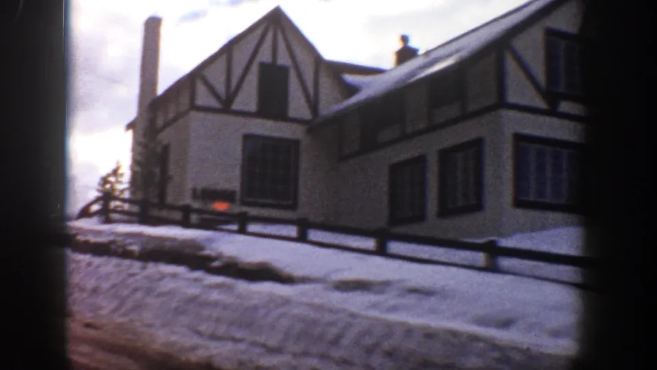 1961 Aspen lodge exterior with Tudor-style gable, chimney, dark window frames, fence, and snow-covered ground.
