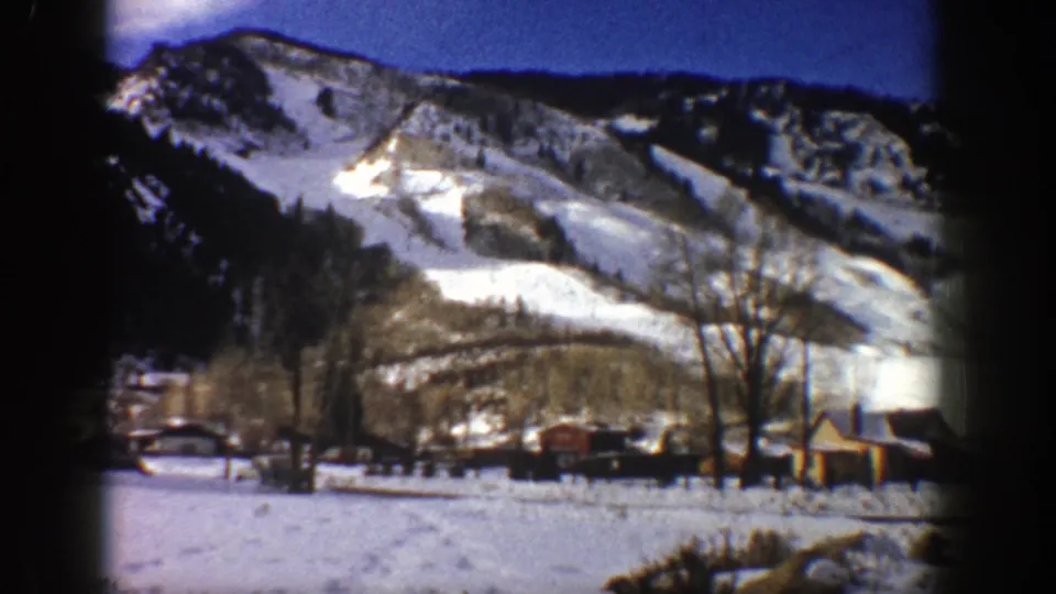 Aspen winter scene on 8mm film, showing ski-cut snowfields, bare trees, small buildings, and open snow below.