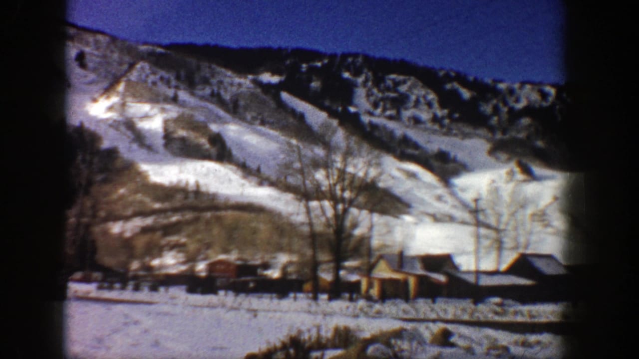 1961 Aspen, Colorado winter view with snow-covered mountains rising behind small cabins, bare trees, and utility poles.