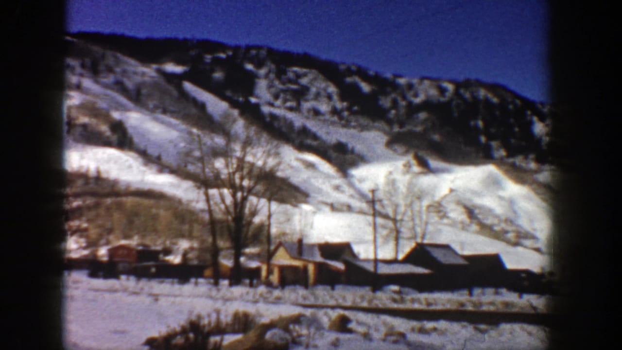Cabins and bare trees sit below bright snowy mountain slopes.