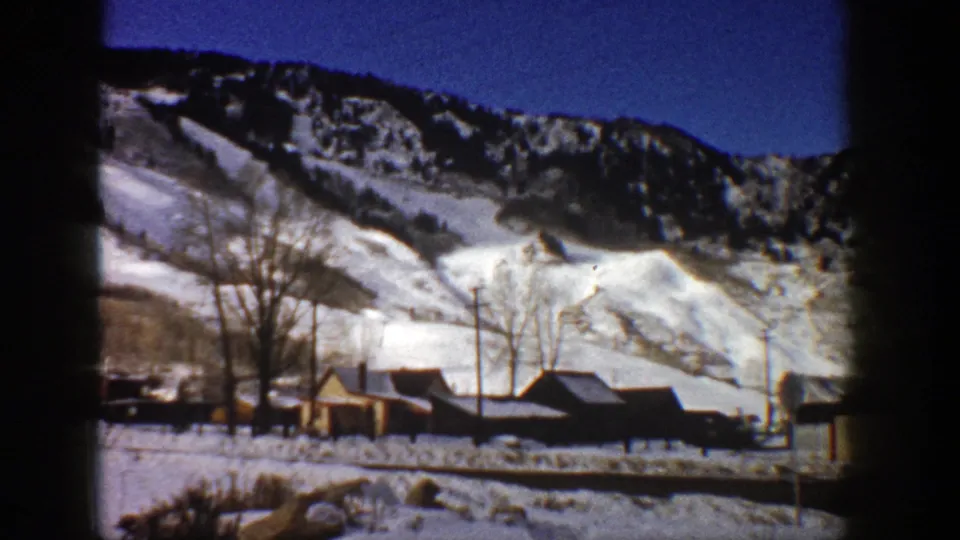 1961 Aspen, Colorado home-movie frame with a snowy mountainside, dark ridge, cabins, bare trees, and roadside foreground.
