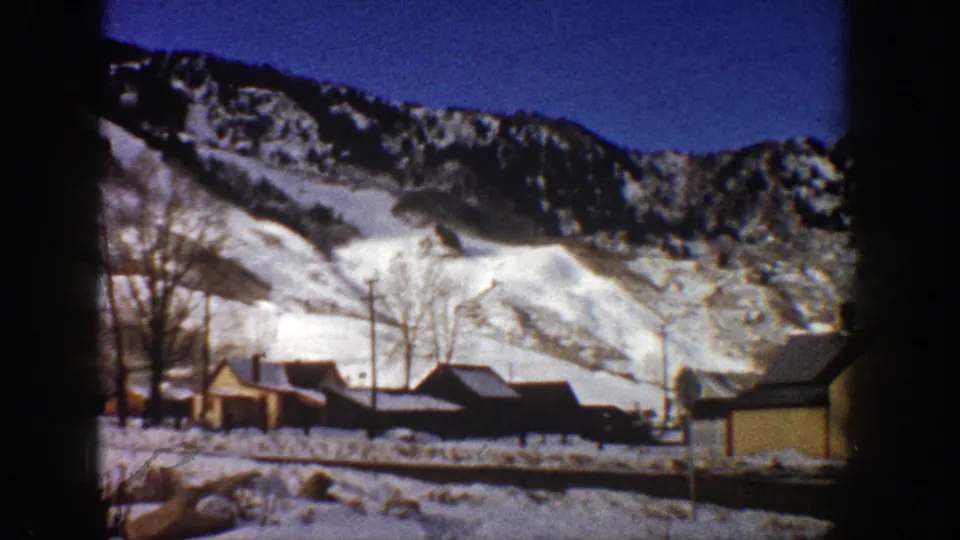 1961 Aspen, Colorado winter frame showing cabins beneath snowy mountains, bare trees, utility poles, and part of a nearby building at right.