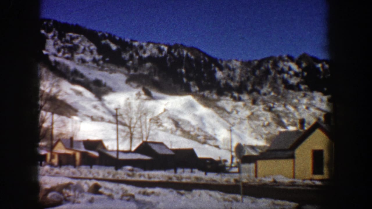 1961 Aspen, Colorado home-movie frame showing snow-covered mountains behind small valley buildings and a yellow house.