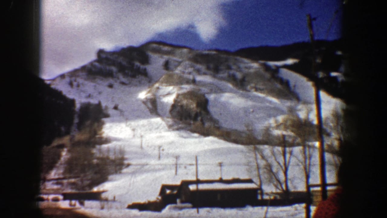 Snowy ski slope with lift poles, bare trees, a small dark building, and rocky mountain ridge.