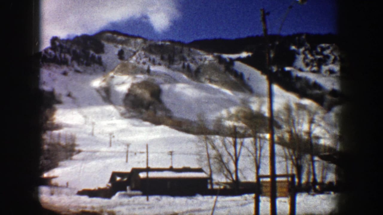 1961 Aspen 8mm frame of a snow-covered ski run with lift poles, bare trees, a small building, and mountain ridge.