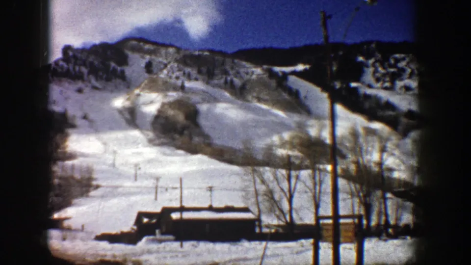 1961 Aspen 8mm frame of a snow-covered ski run with lift poles, bare trees, a small building, and mountain ridge.