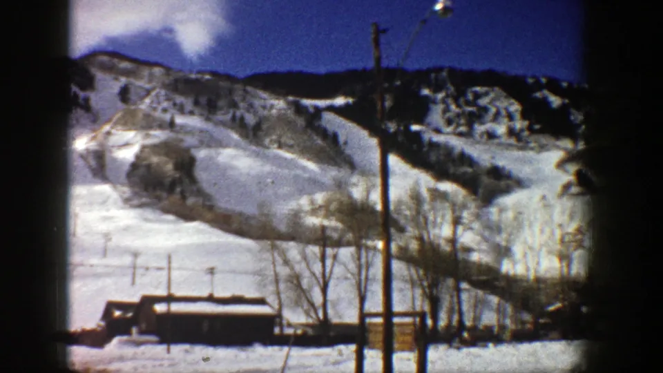 1961 Aspen frame with a snowy slope, central lift pole, bare trees, small building, and darker wooded ridge beyond.