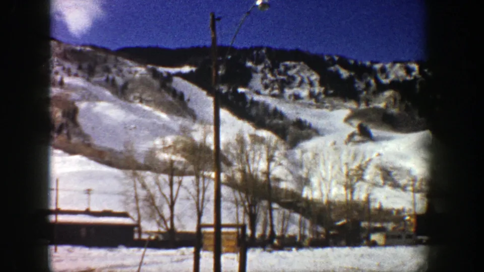 1961 Aspen home-movie frame showing the ski area from a visitor viewpoint, with lift pole, trees, snowfields, and ridge.