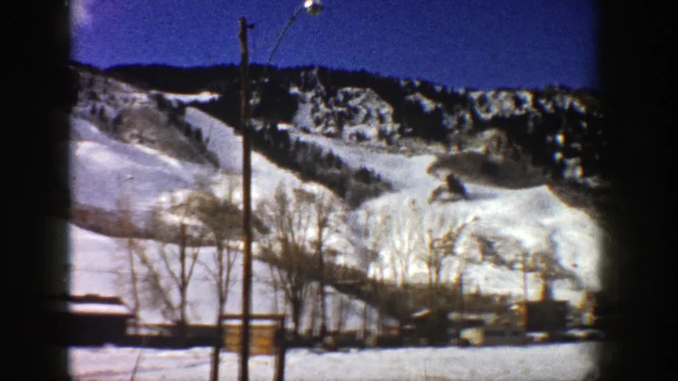 1961 Aspen, Colorado home-movie view of a snow-covered mountain slope with bare trees, utility poles, and low buildings below.