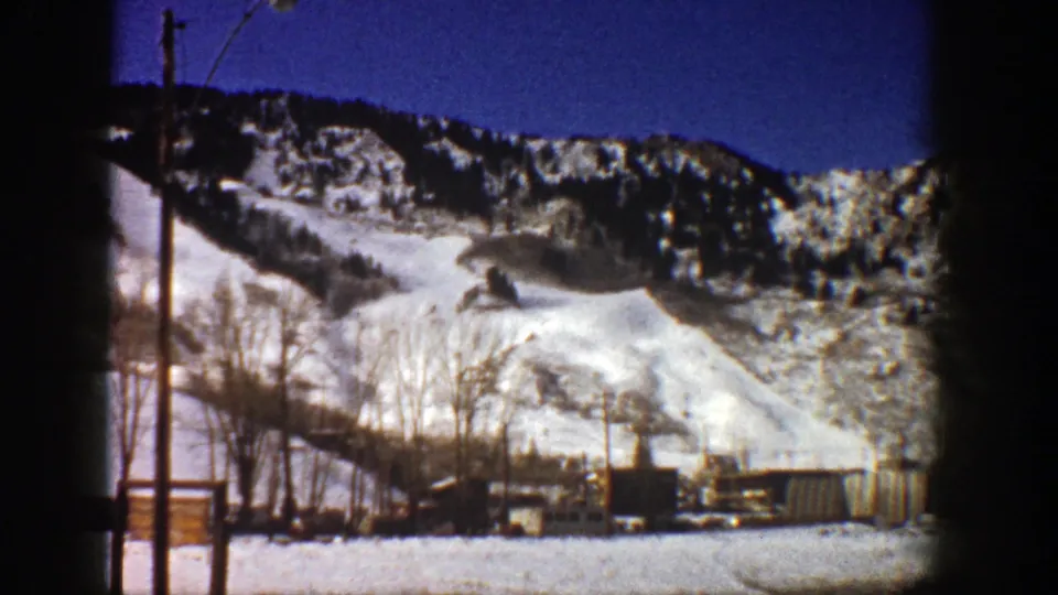 Aspen, Colorado 1961 home-movie frame of a snow-covered mountain run with dark tree lines, poles, and small buildings near the base.