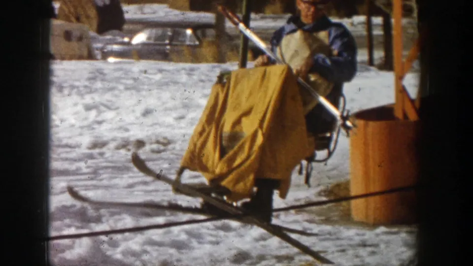 1961 Aspen, Colorado frame showing a person in blue outerwear and glasses seated with skis in a chairlift area on snow.