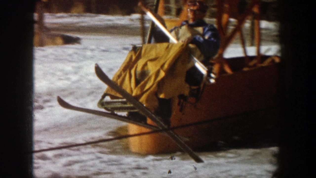 1961 Aspen ski-area view of a worker beside a yellow lift carrier, with skis, poles, snow, and outdoor lift structure visible.