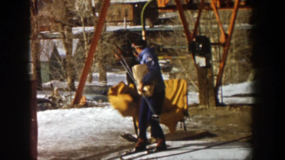 The ski station employee bends beside a yellow carrier in Aspen, with lift supports, skis, snow patches, and bare trees in view.