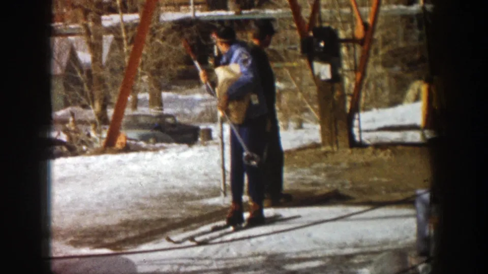 A blue-jacketed worker stands at the Aspen lift station holding ski equipment, with snow, shadows, and orange lift supports nearby.