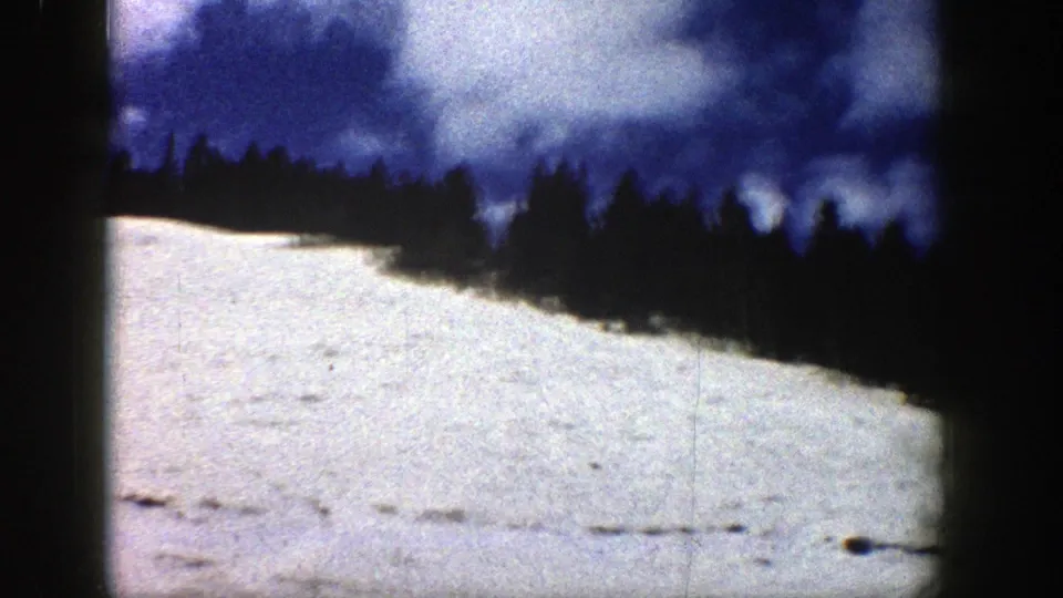 Late frame from the 1961 Aspen clip showing a curving snowy slope, evergreen ridge, blue sky, and surface marks from the 8mm scan.