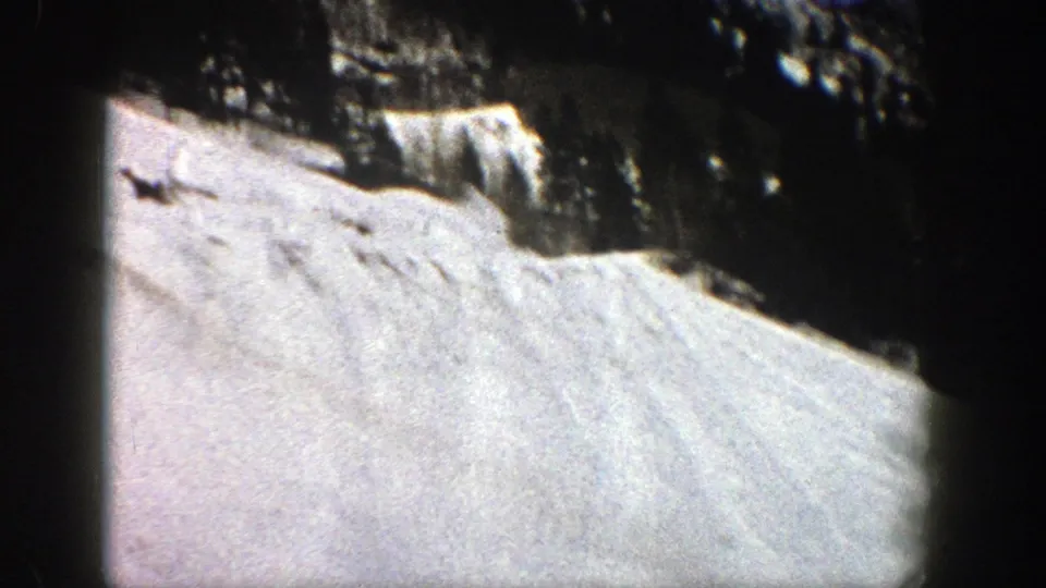 Grainy 8mm view of an Aspen snowfield, with a shadowed evergreen band and rugged rock above the slope.
