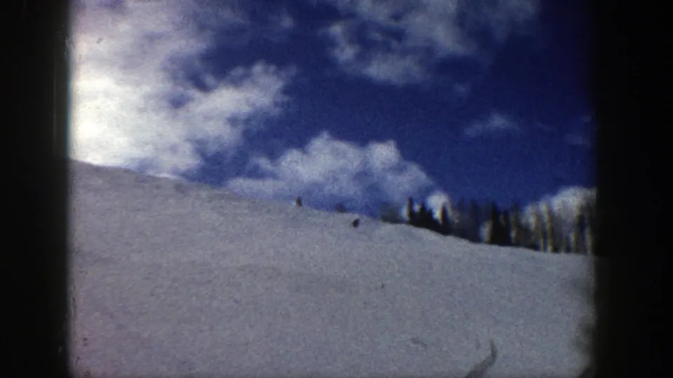1961 Aspen winter frame showing a broad ski slope, scattered clouds, evergreens, and tiny skier figures in the distance.