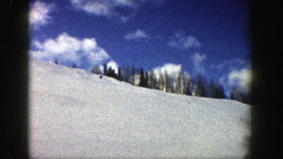 1961 Aspen home-movie frame of a sunlit snow slope with a dark evergreen ridge and blue sky overhead.