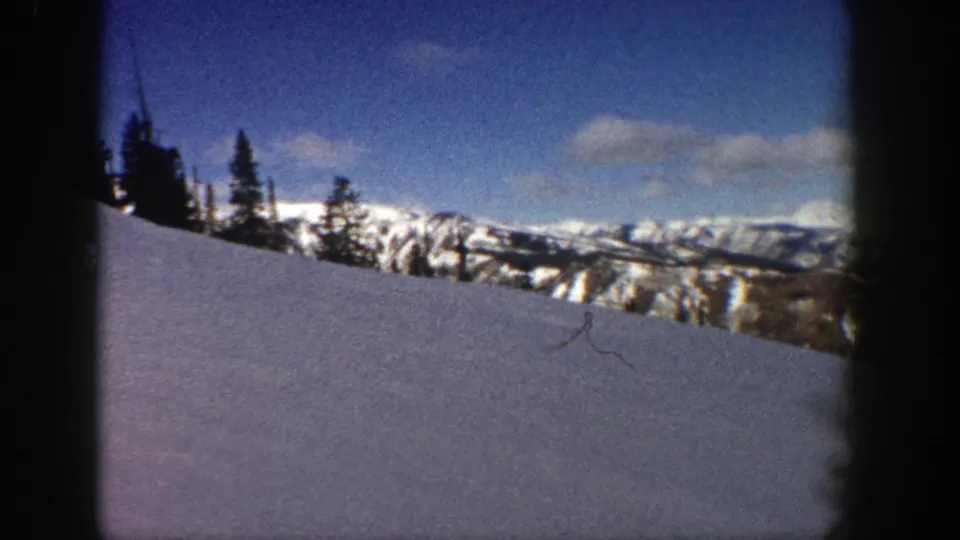 Aspen 8mm frame showing a winter slope, dark trees, distant ridgelines, and scattered clouds across a saturated blue sky.