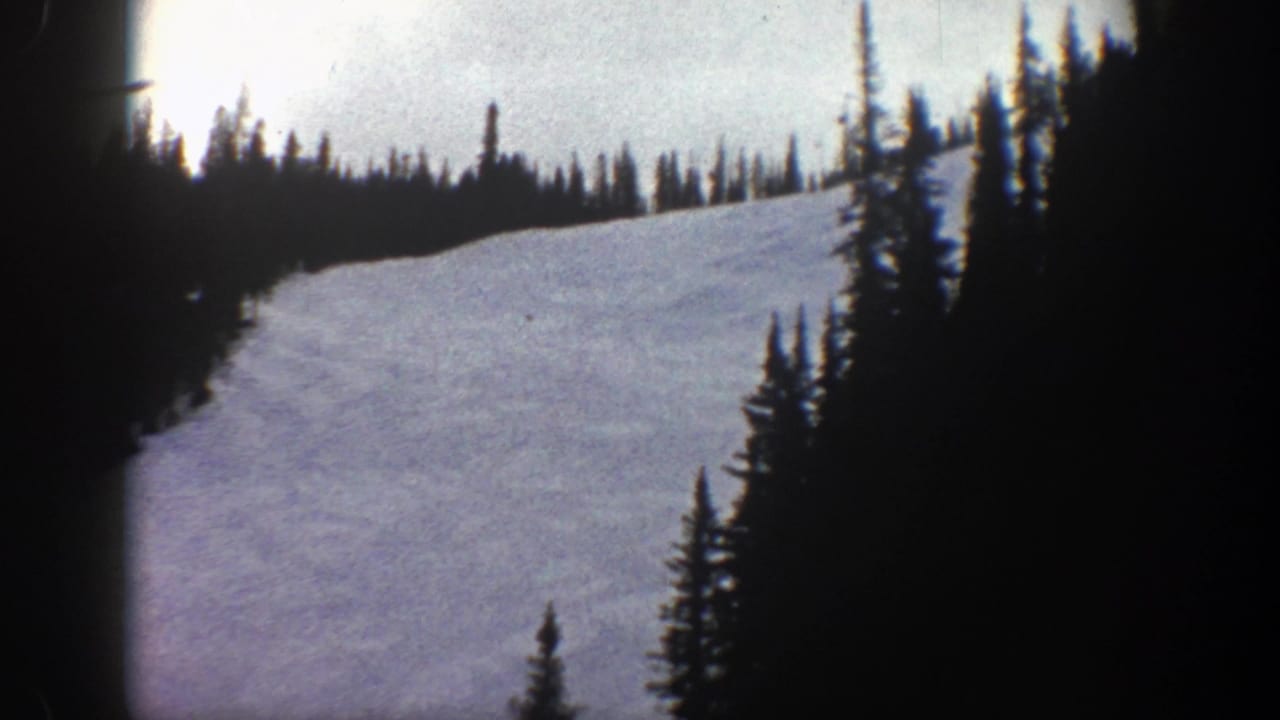 1961 Aspen home-movie frame showing a broad snowy slope framed by evergreen trees and visible 8mm film edges.