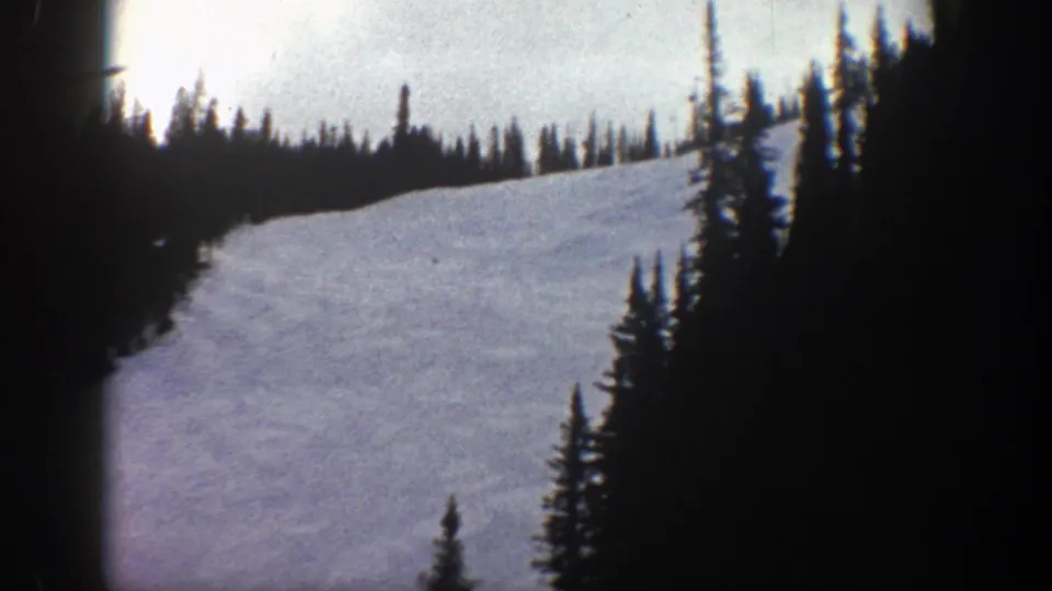 1961 Aspen home-movie frame showing a broad snowy slope framed by evergreen trees and visible 8mm film edges.