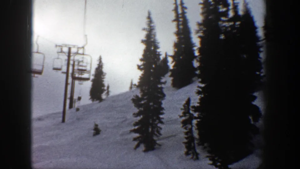1961 Aspen, Colorado 8mm frame showing a chairlift tower, empty chairs, steep snowy ground, and tall conifers along the run.