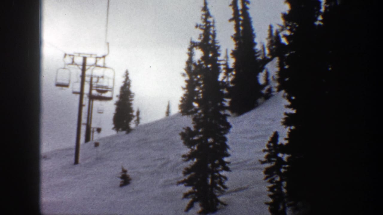 1961 Aspen home-movie frame showing chairlift towers and chairs above a snowy slope with tall evergreens.