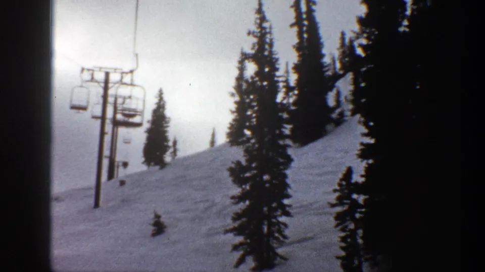 1961 Aspen home-movie frame showing chairlift towers and chairs above a snowy slope with tall evergreens.
