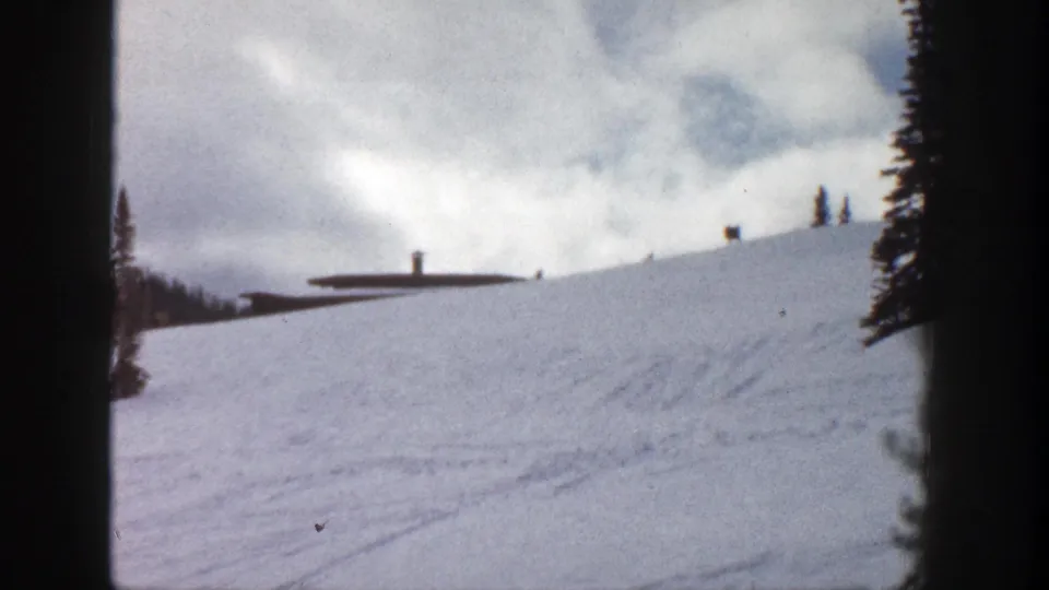 1961 Aspen frame showing a snow-covered ridge, a distant low lodge-like building, scattered conifers, and cloudy winter sky.