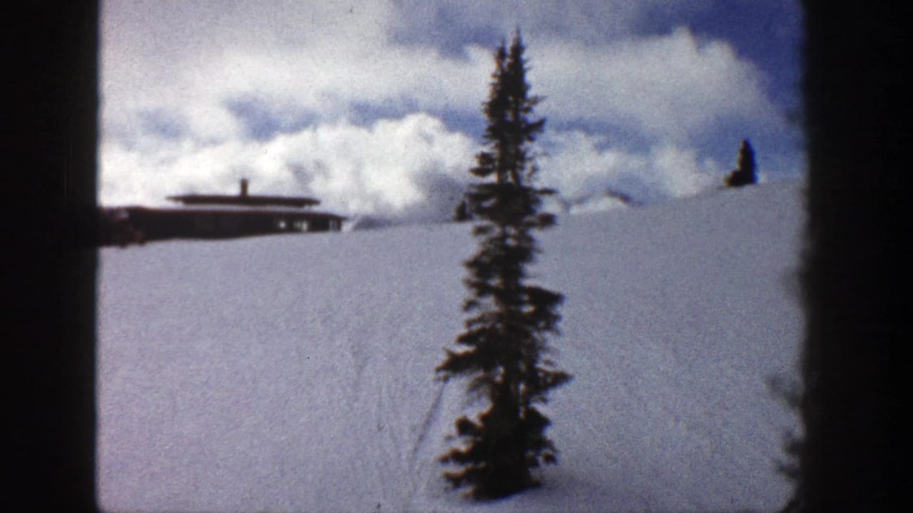 Snowy hillside with a low brown building, evergreen tree, and cloudy sky.