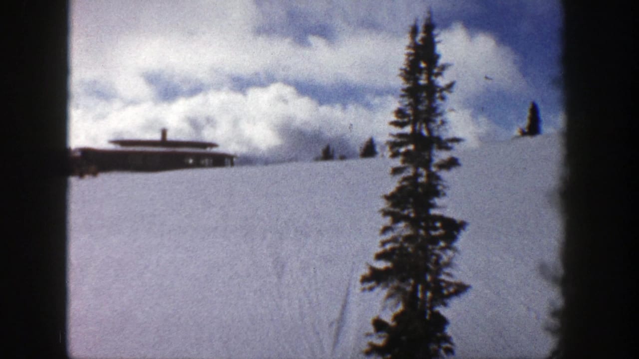 Snow-covered hill with a brown building and evergreen tree under clouds.