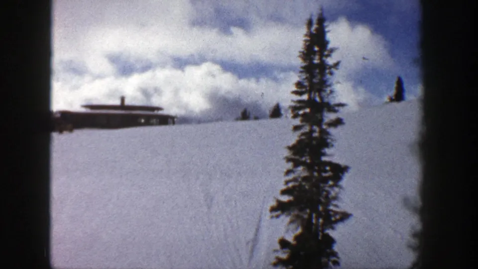 1961 Aspen winter view with a brown flat-roof building on a snowy hill and an evergreen tree in the foreground.