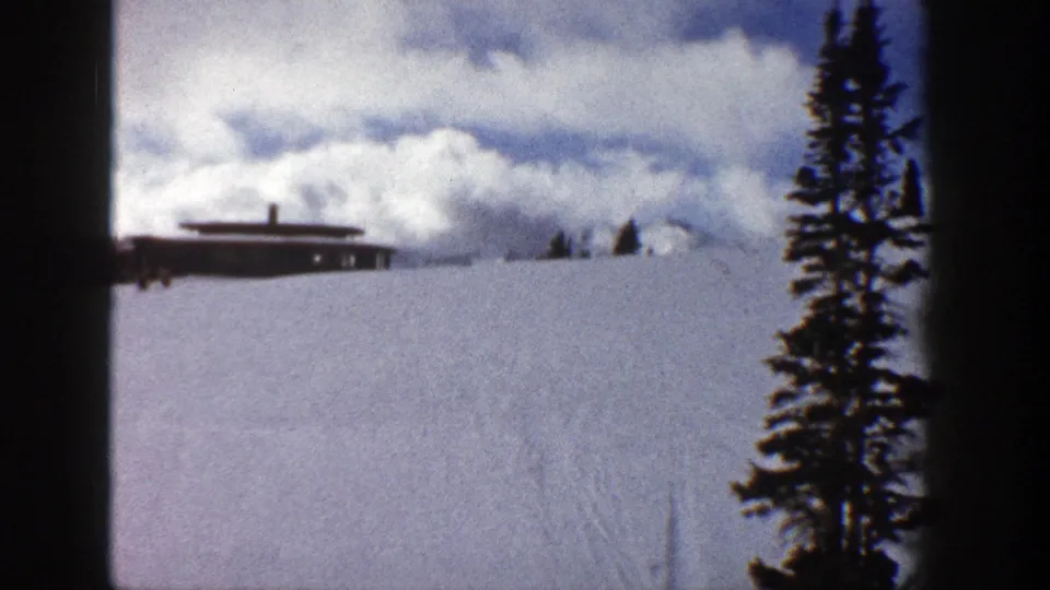 Aspen, Colorado home-movie frame from 1961 showing a distant low building, snowy hillside, clouds, and a foreground evergreen.