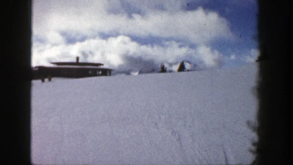 Final 1961 Aspen frame showing the brown building across a broad snowy hill, with clouds and faint tracks in the snow.