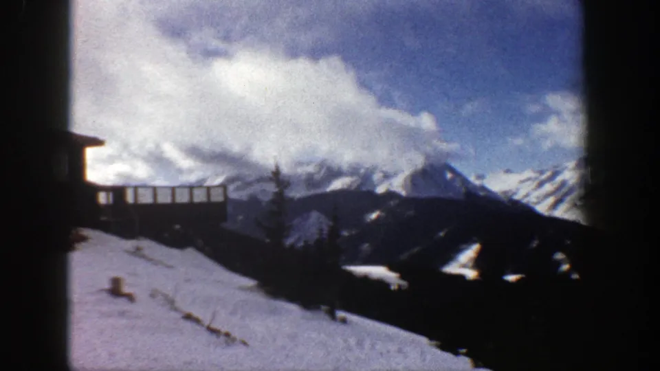 1961 Aspen, Colorado home-movie frame of a snowy slope near a wooden structure, with evergreen ridges and clouded peaks.