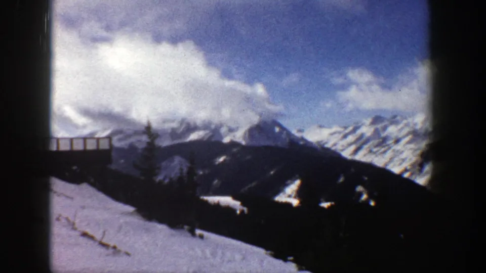 1961 Aspen, Colorado frame showing a snowy mountain panorama, partial wooden railing, evergreens, and bright cloud cover.