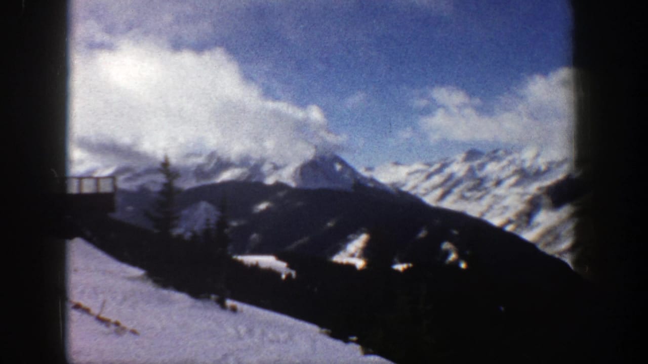 Snowy mountain ridge under clouds with a dark slope and small structure at the far left.