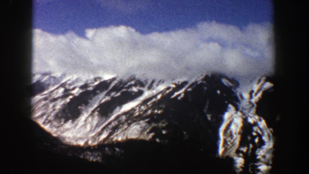 1961 Aspen, Colorado 8mm frame showing distant snow-covered mountain ridges under a blue sky with white clouds, bordered by dark film edges.