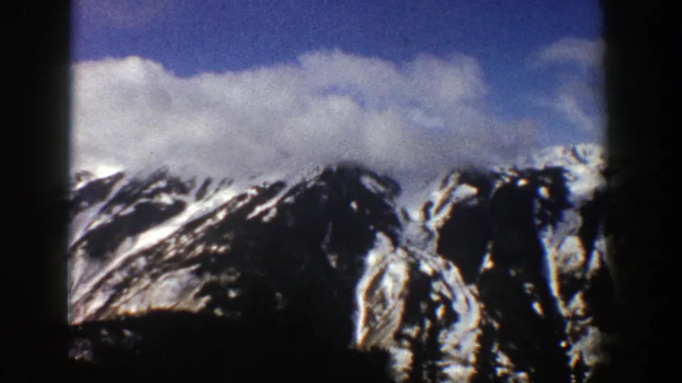 1961 Aspen, Colorado 8mm frame showing a distant mountain range with snow patches, dark rocky slopes, thick white clouds, and visible film-frame borders.