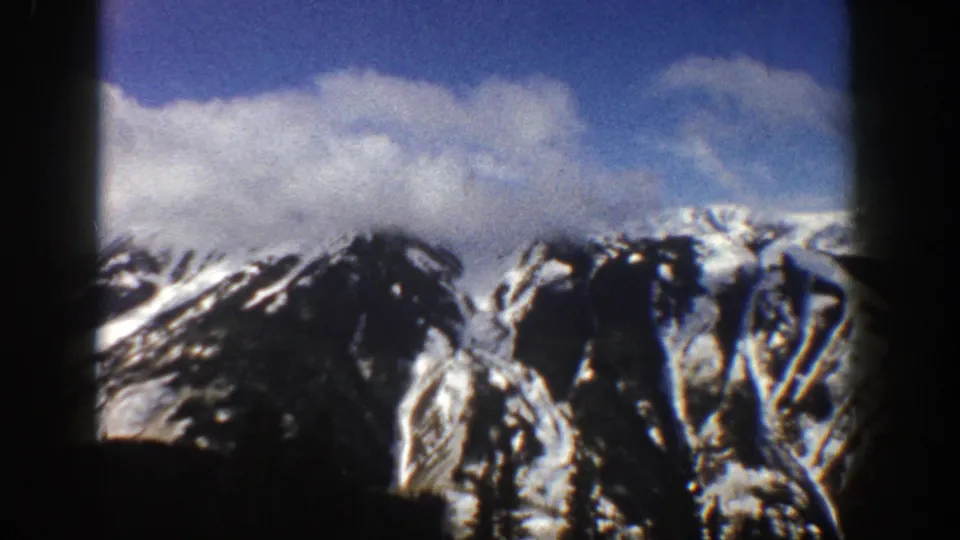 1961 Aspen, Colorado 8mm frame with snow-covered mountain peaks and rocky faces beneath a cloud-filled blue sky, partly framed by dark vertical borders.