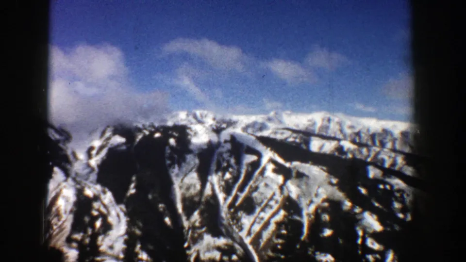 1961 Aspen, Colorado 8mm frame with snow-striped mountain peaks, blue sky, clouds, and visible dark frame edges.