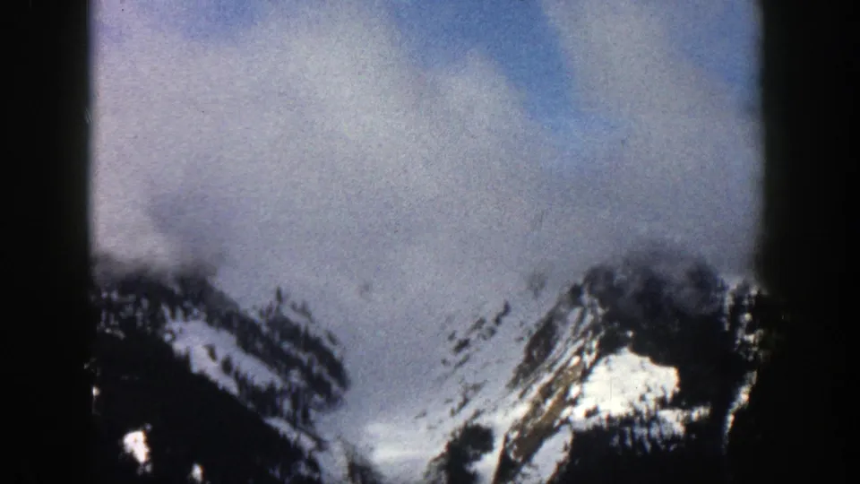 1961 Aspen, Colorado 8mm frame showing snow-striped alpine peaks partly veiled by clouds under a blue-gray sky.