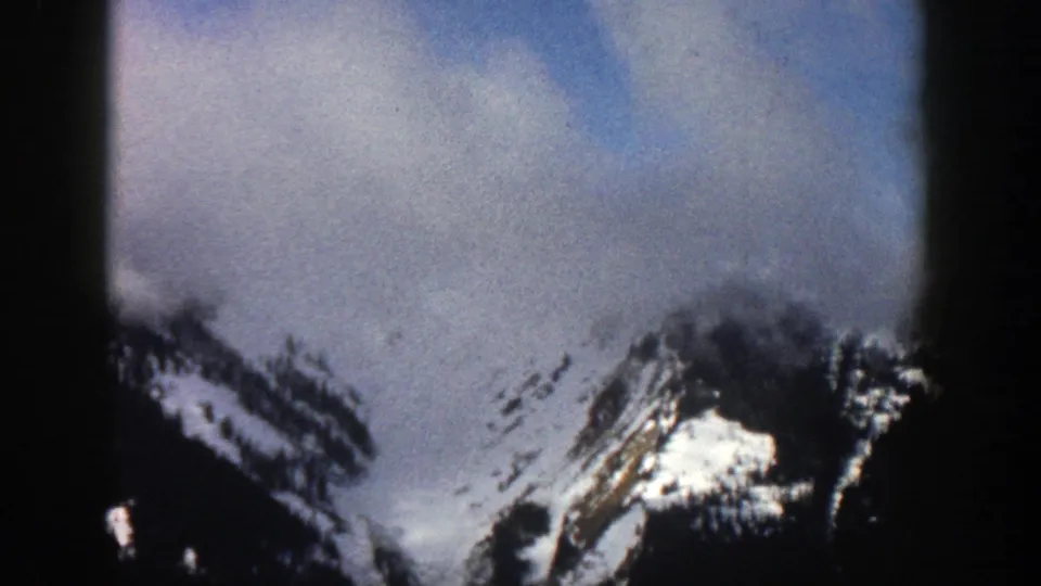 1961 Aspen, Colorado home-movie frame with softened snowy peaks, cloud cover, and blue-toned 8mm color fading.